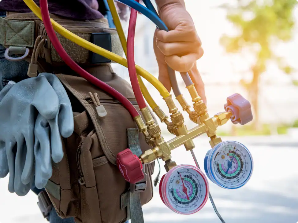 A person holds HVAC gauges with colored hoses; gloves and a tool bag are also visible, suggesting air conditioning or refrigeration system maintenance work.